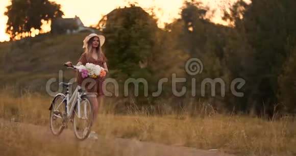 年轻漂亮的金发女孩穿着连衣裙戴着帽子骑着自行车和花慢慢地走在路上视频的预览图