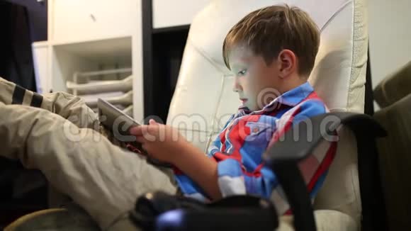 快乐的小男孩坐在家里的椅子上玩他的平板电脑视频的预览图