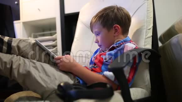 快乐的小男孩坐在家里的椅子上玩他的平板电脑视频的预览图