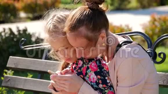 年轻的母亲带着女儿在户外放松他们坐在长凳上玩公园里的智能手机一个年轻人视频的预览图