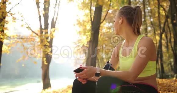 公园里的女跑步者穿着慢跑服的可爱的年轻女子在跑步后休息听音乐视频的预览图