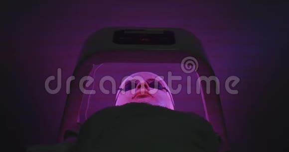 带着面具和眼镜的女人躺在水疗沙龙的光机下面部LED光疗Cosmetology和视频的预览图