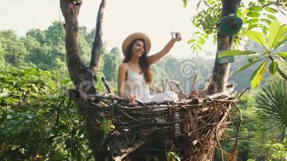 年轻的快乐混血旅游女孩穿着白色连衣裙用手机拍摄自拍照片坐在装饰草窝里视频的预览图