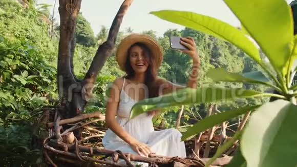 年轻的快乐混血旅游女孩穿着白色连衣裙用手机拍摄自拍照片坐在装饰草窝里视频的预览图