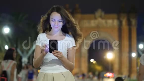 晚上年轻女子在巴塞罗那的地标上浏览智能手机视频的预览图