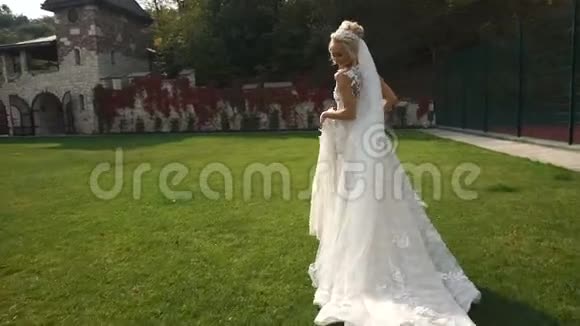 年轻迷人的金发新娘穿着美丽的长裙沿着盛开的花园奔跑视频的预览图