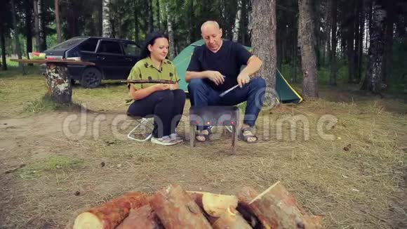 在帐篷附近一个女人用鱼把烤架翻过来男人在烤架里搅动煤视频的预览图