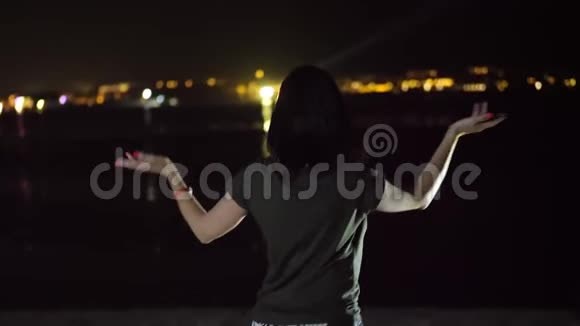 夜晚一个女人在跳舞从后面看在水面上发光的城市的灯光下视频的预览图