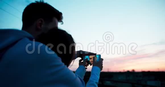 后观幸福微笑的情侣在手机上看照片在屋顶欣赏日落视频的预览图