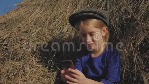 年轻可爱的女孩带着帽子玩得很开心坐在干草堆里用电话坐在干草上的小女孩视频的预览图