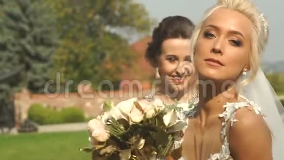 幸福美丽的金发新娘迷人的微笑和婚礼花束正在与她的伴娘在花园里摆姿势视频的预览图
