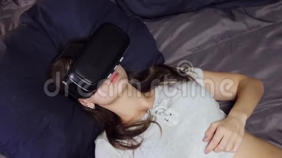 年轻女性使用虚拟现实的俯视图观看躺在床上枕头上的VR视频视频的预览图