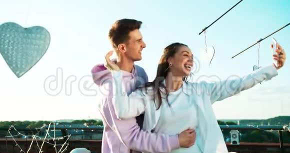 这对年轻迷人的情侣在屋顶上通过手机笑着自拍的侧面画像视频的预览图