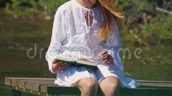 一个穿着白色连衣裙的年轻女子坐在码头上的河边读一本书视频的预览图