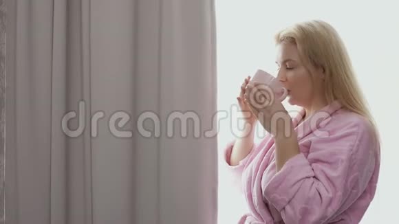 清晨年轻的白种人金发碧眼喝着绿茶站在窗边视频的预览图