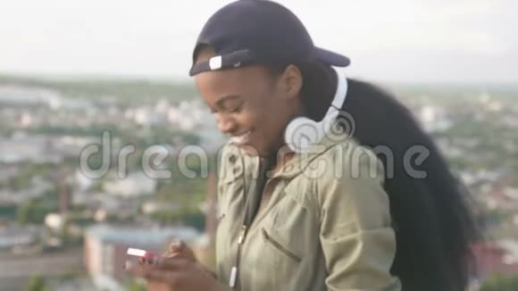 戴帽子的美国黑人女孩戴着耳机听音乐微笑着模糊的城市景观背景视频的预览图