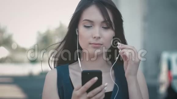 使用智能手机和在户外街头背景下听耳机音乐的年轻美女画像视频的预览图