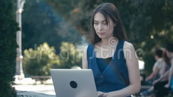 在城市街道背景长凳上的公园里用笔记本电脑工作的年轻女子视频的预览图
