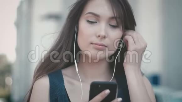 使用智能手机和在户外街头背景下听耳机音乐的年轻美女画像视频的预览图