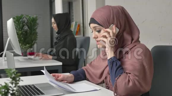 年轻漂亮的女孩穿着粉红色的头巾拿着文件和手机说话办公室里的阿拉伯妇女60英尺视频的预览图