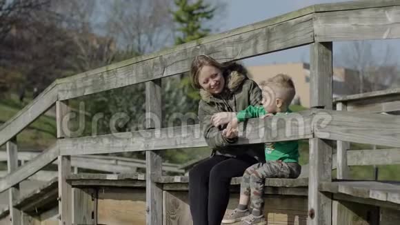 快乐的年轻妈妈和她的小儿子坐在木桥上把石头扔进水里视频的预览图