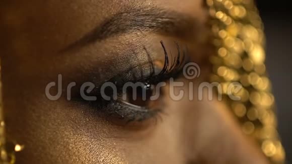 张开紧闭的眼睛用青铜眼影和猫眼盯着戴着金色珠宝的非洲女孩的脸视频的预览图