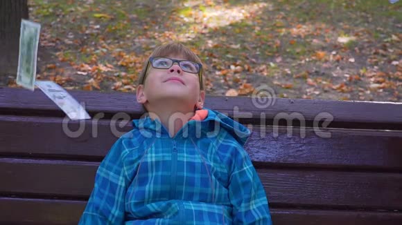 一个可爱的小孩坐在公园的长椅上一张从天上掉下来的钞票的肖像视频的预览图