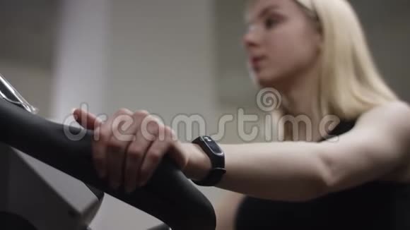 金发女孩骑着一辆运动自行车转动踏板她在新健身房里看其他运动员视频的预览图
