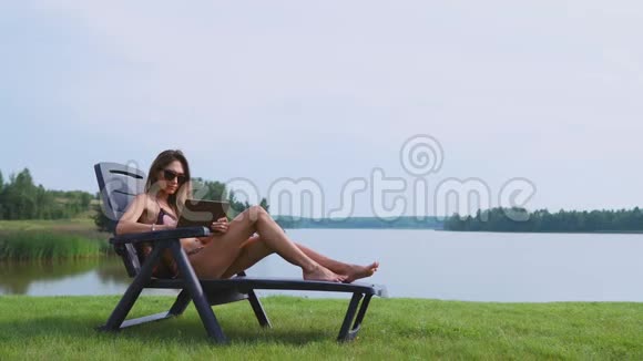 穿着泳衣戴着平板电脑的女人躺在阳光休息室里手指触摸着平板电脑的屏幕微笑着视频的预览图