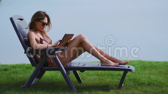 美丽的黑发女郎穿着泳衣躺在湖上的日光休息室里微笑着通过互联网远程工作视频的预览图