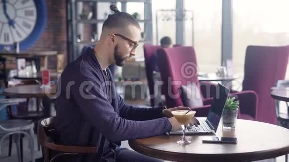一个穿着黑色T恤和蓝色夹克的男人用笔记本电脑坐在桌子旁的侧面年轻的时髦女郎视频的预览图