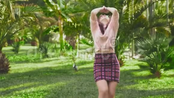 年轻漂亮的红发女孩在公园里跳舞穿迷你裙的女人在热带公园的背景下跳舞视频的预览图