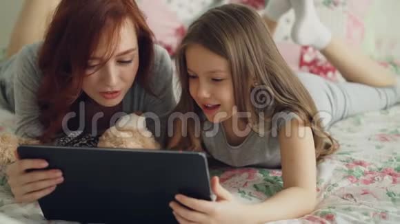 一位微笑可爱的女孩和年轻的母亲穿着睡衣躺在床上笑着看着数码平板电脑视频的预览图