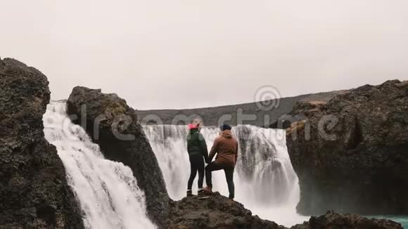 年轻夫妇站在岩石上看着冰岛一个强大的瀑布手牵着手视频的预览图