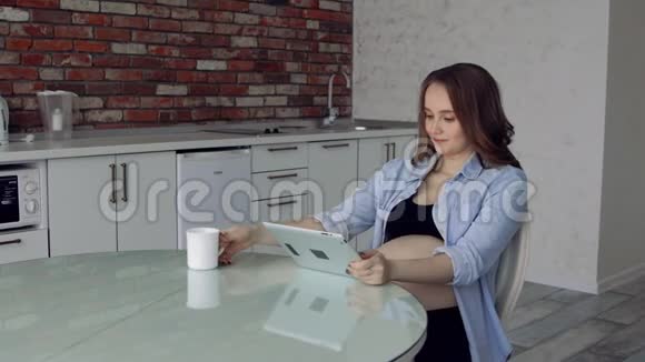 幸福的孕妇坐在玻璃餐桌旁喝咖啡用平板电脑网上购物视频的预览图