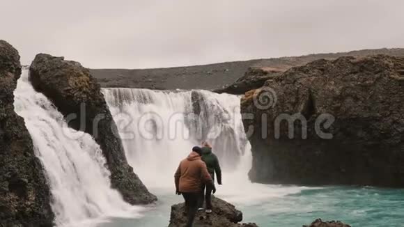 年轻夫妇在冰岛徒步旅行旅行男女站在强大的瀑布附近举手视频的预览图