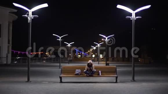 快乐的女孩在玩手机应用程序上的小玩意坐在夜间城市的长凳上视频的预览图