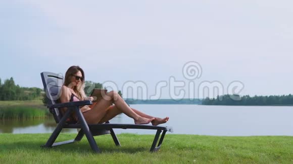 美丽的黑发女郎穿着泳衣躺在湖上的日光休息室里微笑着通过互联网远程工作视频的预览图