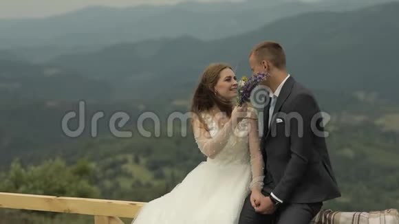 新郎和新娘坐在山岗附近的篱笆上新婚夫妇家庭视频的预览图