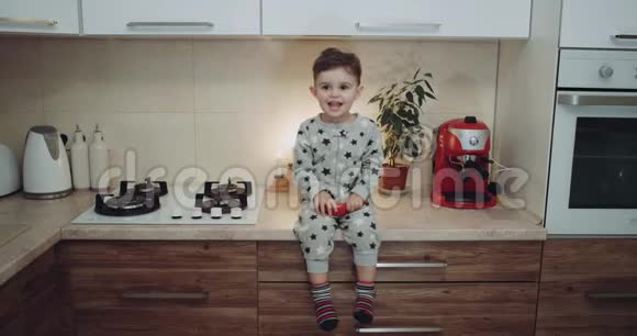 穿着睡衣的小男孩坐在厨房的桌子上快乐地拍手用手亲吻4k视频的预览图