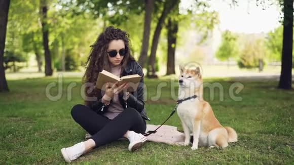 有魅力的年轻女子正坐在公园的草地上看书而她养得很好的狗正坐在她的主人旁边视频的预览图