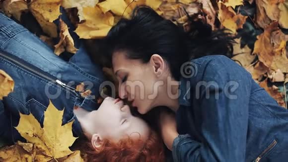 一位美丽的母亲和她可爱的女儿躺在秋天公园的一片黄叶上妈妈吻女儿幸福的家庭视频的预览图