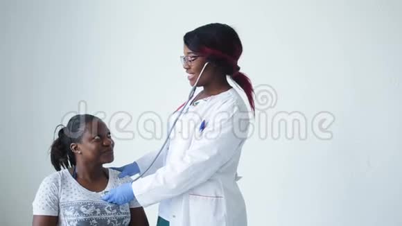 一位年轻微笑的非洲医生正在用听诊器在白色上方倾听女性病人的呼吸视频的预览图