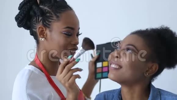 快乐的非洲化妆师正用粉涂在令人惊叹的非洲模特的脸上带着美丽的微笑视频的预览图
