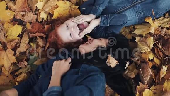 一位美丽的母亲和她可爱的女儿躺在秋天公园的一片黄叶上妈妈吻女儿幸福的家庭视频的预览图
