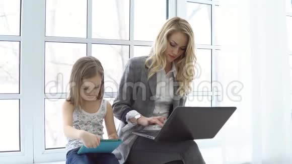 商务女性带着笔记本电脑小女孩带着平板电脑坐在大窗户旁视频的预览图