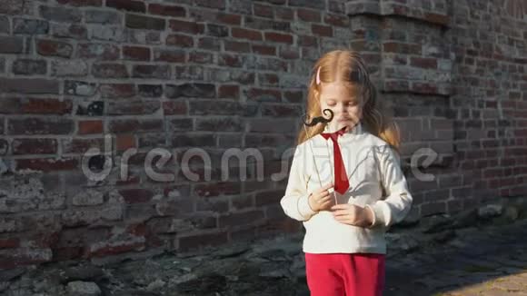 小女孩拿着一根纸胡子在棍子上站在砖墙旁边视频的预览图