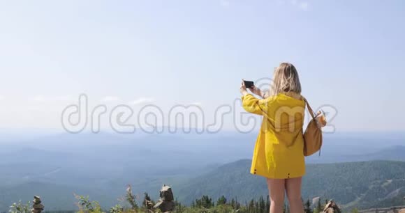 穿着黄色雨衣的女孩在智能手机上拍照站在山顶蓝天和群山上视频的预览图