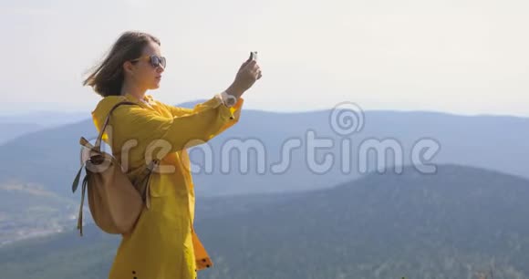 穿着黄色雨衣的女孩在智能手机上拍照站在山顶蓝天和群山上视频的预览图
