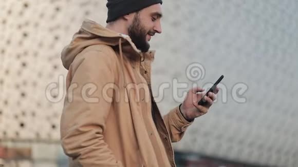 快乐的年轻胡须男用智能手机在购物中心附近的街道上行走他穿着一件秋装视频的预览图